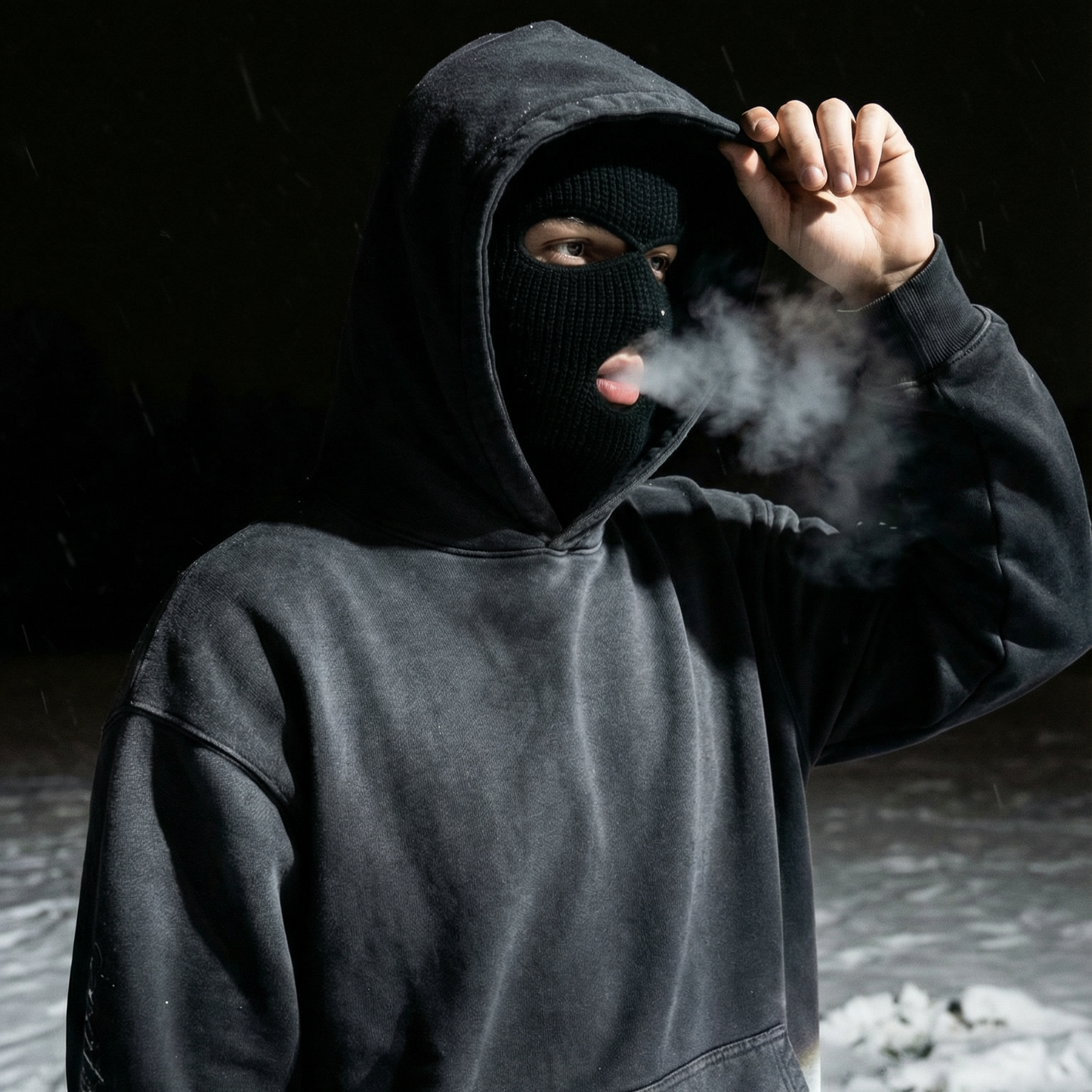 Person wearing a dark hoodie and balaclava exhaling steam in a snowy landscape